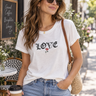 A woman wearing a white graphic t-shirt with the word 'LOVE' printed in black gothic letters and a small red rose, paired with light blue jeans and sunglasses, standing on a sunny street.