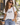 A woman wearing a white graphic t-shirt with the word 'LOVE' printed in black gothic letters and a small red rose, paired with light blue jeans and sunglasses, standing on a sunny street.