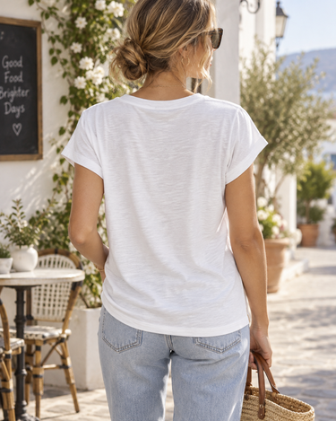 A woman viewed from behind wearing a white short-sleeve crewneck t-shirt and light blue jeans, standing on a sunny outdoor patio walkway.