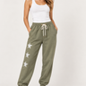 A woman modeling olive green jogger sweatpants with white embroidered stars down one leg, styled with a white tank top and white sneakers against a light gray studio background.