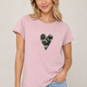 A woman wearing a light pink heathered t-shirt with a green camouflage heart graphic on the chest, paired with light blue jeans, against a neutral beige background.