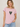 A woman wearing a light pink heathered t-shirt with a green camouflage heart graphic on the chest, paired with light blue jeans, against a neutral beige background.