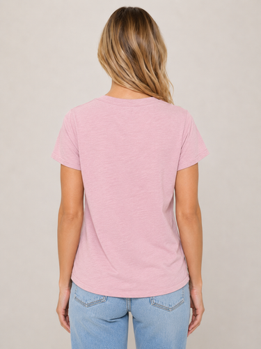 A woman viewed from behind wearing a light pink short-sleeve crewneck t-shirt and light blue jeans, standing against a neutral beige studio background.
