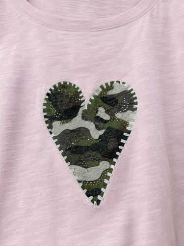 A close-up of a light pink heathered t-shirt featuring a central heart-shaped patch with a green, black, and gray camouflage pattern and a white serrated border.