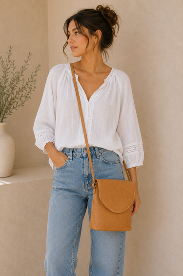 Woman wearing a white blouse and blue jeans, holding a brown leather bag against a beige wall.