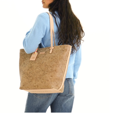 A woman wearing a light blue sweater and jeans carries a large tan embossed leather tote bag with beige handles and trim against a plain white background.