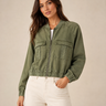 A woman wearing an olive green cropped utility jacket with a front zipper and flap chest pockets, paired with white pants, against a plain beige studio background.
