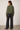 A woman shown from the back wearing an olive green cropped jacket and dark charcoal wide-leg jeans, standing against a plain light gray studio wall.
