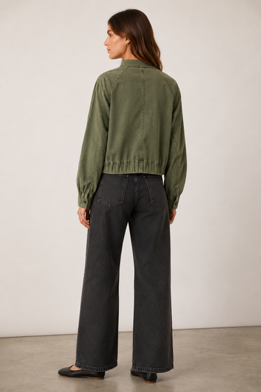 A woman shown from the back wearing an olive green cropped jacket and dark charcoal wide-leg jeans, standing against a plain light gray studio wall.
