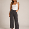 A woman modeling a pair of dark gray wide-leg cropped jeans with a white ribbed tank top and black strappy heels, standing against a neutral beige studio background.