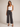 A woman modeling a pair of dark gray wide-leg cropped jeans with a white ribbed tank top and black strappy heels, standing against a neutral beige studio background.