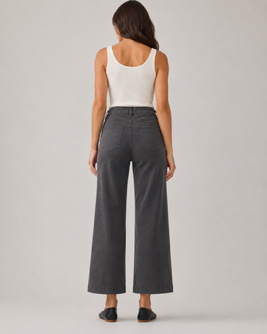A woman viewed from the back wearing dark charcoal gray wide-leg cropped pants, a white ribbed tank top, and black flats, standing against a light gray studio background.