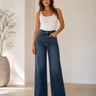 A woman modeling a pair of high-waisted, wide-leg blue jeans with a white tank top, standing in a minimalist room with a beige floor and white walls.