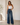 A woman modeling a pair of high-waisted, wide-leg blue jeans with a white tank top, standing in a minimalist room with a beige floor and white walls.