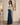 A woman stands in a boutique wearing a fitted cream long-sleeve top and high-waisted, wide-leg dark blue denim jeans. The background features a warm neutral interior with a large floor-to-ceiling mirror on the left and a beige ceramic vase on a pedestal to the right.