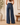 A woman shown from the back wearing high-waisted, wide-leg blue denim jeans with a brown leather patch at the waistband, standing in a warm, neutral-toned room.