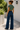 A woman stands outdoors in a sunlit alley wearing a cropped sage green t-shirt and high-waisted, wide-leg dark blue jeans, viewed from behind.