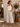 A woman stands in a boutique wearing a white sleeveless tank top and light beige wide-leg cropped pants, with brown flat sandals. A clothing rack, mirror, wooden table with a vase of greenery, and a woven basket are visible in the softly lit interior.