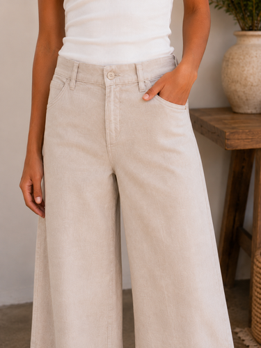 A person wearing light beige wide-leg pants and a white ribbed tank top, standing indoors with one hand in a pocket. A wooden side table and ceramic vase are visible in the softly blurred background.