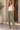A woman stands in a sunlit entryway wearing olive green wide-leg pants, a white ribbed tank top, and taupe loafers, with her hands in the pockets of the pants.