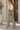 A woman wearing olive green wide-leg pants, a white sleeveless top, and beige flat shoes, standing on a tiled porch.