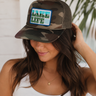 A woman wearing a camouflage trucker hat with a rectangular front patch that reads 'LAKE LIFE' in a blue and green graphic design. She is smiling and posing indoors with a white wall and green plant leaves in the background.