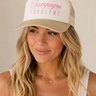 A woman wearing a beige and white trucker hat with pink text that reads 'Champagne PROBLEMS' against a warm beige background.