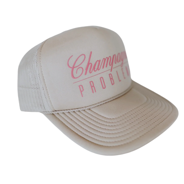 A beige trucker hat with a pink embroidered graphic on the front panel that reads 'Champagne PROBLEM'.