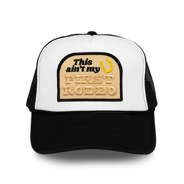 A black and white trucker hat with a beige embroidered patch on the front that reads 'This ain't my FIRST RODEO' with a yellow horseshoe graphic, shown on a white background.