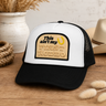 A black and white trucker hat with a tan embroidered patch reading 'This ain't my first rodeo' and a yellow horseshoe, styled on a wooden surface with rustic decor.