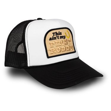 A black and white trucker hat with a beige embroidered patch on the front that reads 'This ain't my FIRST RODEO' with a small yellow riding crop accent, shown on a white background.