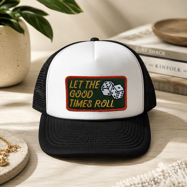 A black and white trucker hat with a green and gold embroidered patch reading 'LET THE GOOD TIMES ROLL' and showing two dice, styled on a light wood tabletop with books and a plant in the background.