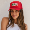 A woman wearing a red trucker-style cap with a white front patch that reads 'Spicy' in green script with a red chili pepper graphic, posed against a light textured wall.