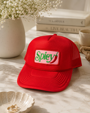 A red trucker-style cap with a front patch reading 'Spicy' in green over a pink background, styled on a marble surface with flowers, books, and a cup nearby.