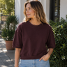 A woman wearing a burgundy sweatshirt with 3/4 sleeves and blue jeans outside a shop.