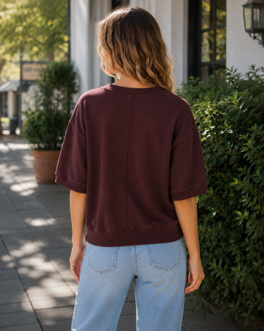 A woman wearing a burgundy sweatshirt with 3/4 sleeves and blue jeans outside a shop.