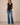 A woman modeling a sleeveless navy blue draped wrap-style top with blue flare jeans, standing against a plain beige studio backdrop.