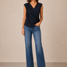 A woman modeling a sleeveless navy blue draped wrap-style top with blue flare jeans, standing against a plain beige studio backdrop.