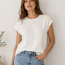 A woman wearing a white short-sleeve crewneck top and light blue jeans, standing against a plain off-white wall.