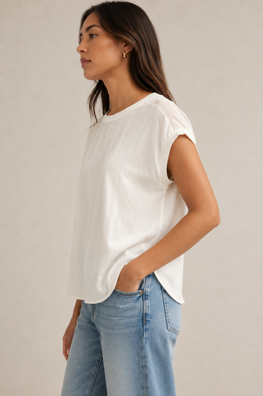 A woman wearing a white short-sleeve crewneck top and light blue jeans, shown in a side-profile view against a plain light beige studio background.