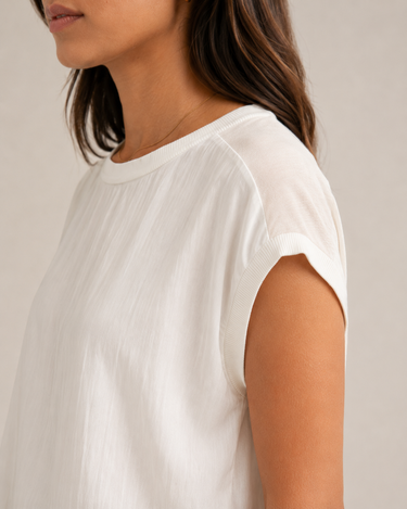 Close-up side view of a woman wearing a white short-sleeve top against a plain light beige background.