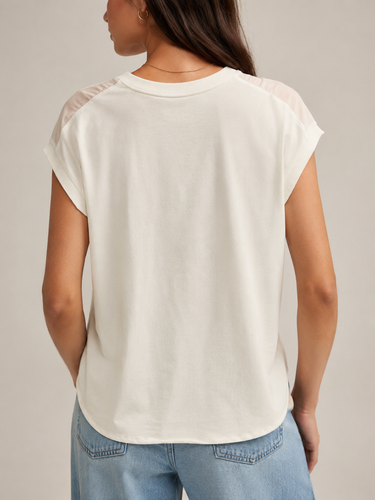 A woman shown from the back wearing a short-sleeve cream-colored top with sheer shoulder panels and light blue jeans, set against a plain light gray studio background.