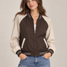 a woman wearing a reversible baseball brown jacket with black jeans on a neutral background.