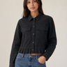 A woman modeling a black cropped denim jacket with a button-down front, chest flap pockets, and a ruffled hem, paired with blue jeans against a plain light beige studio background.