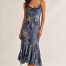 A woman modeling a blue floral satin slip dress with thin straps and a ruffled hem, standing against a plain light beige studio background.