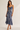 A woman modeling a blue floral satin slip dress with thin straps and a ruffled hem, standing against a plain light beige studio background.