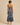 A woman viewed from the back wearing a sleeveless satin slip dress with thin straps and a blue-gray watercolor floral print with gold accents, paired with beige heels, against a solid beige studio background.