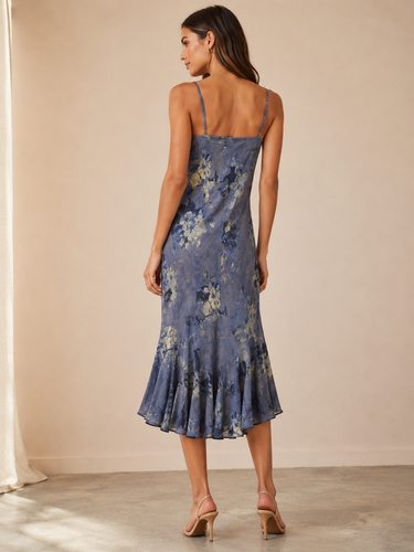 A woman modeling a blue floral midi slip dress with spaghetti straps and a ruffled hem, shown from the back in a warm beige studio setting.