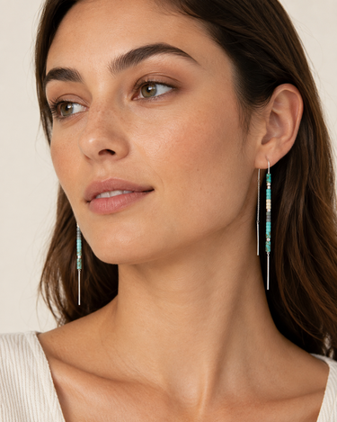 A close-up of a woman wearing long beaded drop earrings with turquoise, white, and gold beads on silver-toned wires, against a soft beige background.