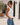 A woman wearing a fitted white sleeveless tank top and light blue jeans, standing in a sunlit courtyard with white walls, arched windows, and a potted olive tree.